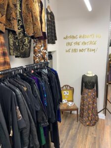 photo of the inside of a shop, a rail full of dark clothes is on the left on the white wall in front is a gold decal that reads "nothing haunts you like the vintage you didn't buy"