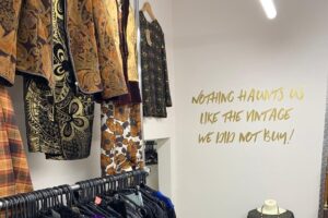 photo of the inside of a shop, a rail full of dark clothes is on the left on the white wall in front is a gold decal that reads "nothing haunts you like the vintage you didn't buy"