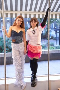 Actresses Mia Tomlinson and Isabella Pappas pose in FARA Charity Shop window in outfits made of recycled clothing