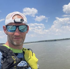 Man wearing mirrored sunglasses and white backwards-facing baseball cap, looks into camera, with backdrop of River Danube