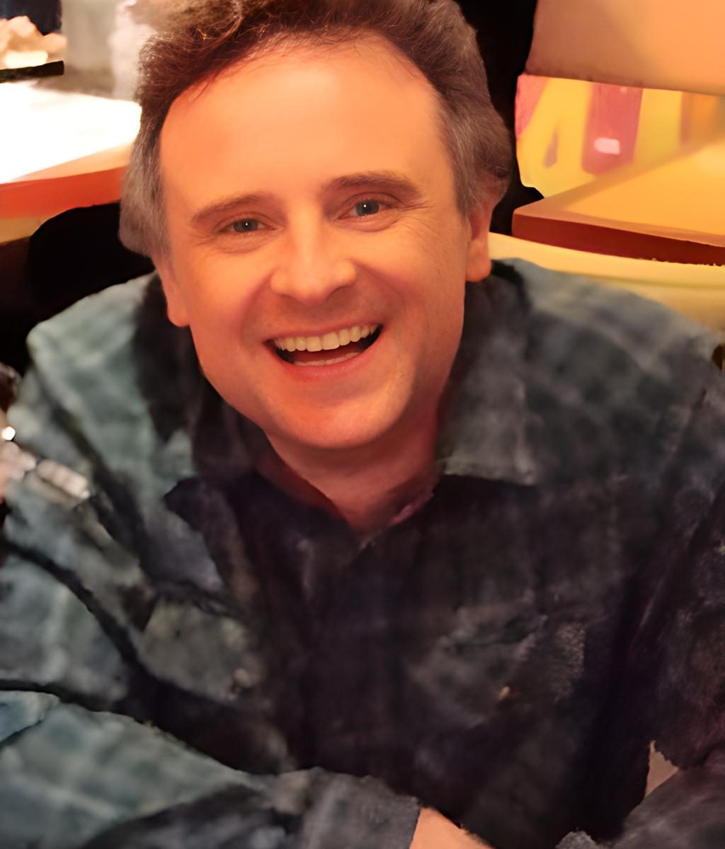 A middle aged white man with brown hair smiles at the camera. He's wearing a dark blue checked shirt