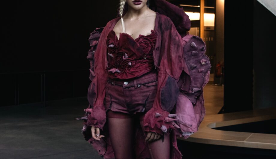 A photo of a London College of Fashion student wearing an outfit made from unsellable denim. The outfit is dyed pink and includes, shorts, a corset and a large jacket. The student has a blonde braid falling over their face