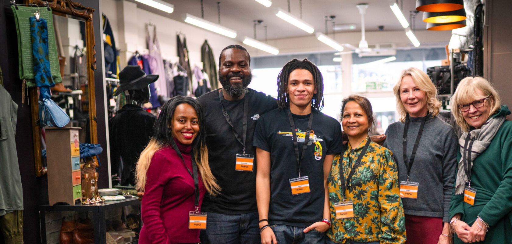 ChatGPT said: A group of six FARA staff and volunteers stand together inside a brightly lit charity shop, smiling at the camera. They are wearing name badges and lanyards, surrounded by neatly displayed clothing and accessories.