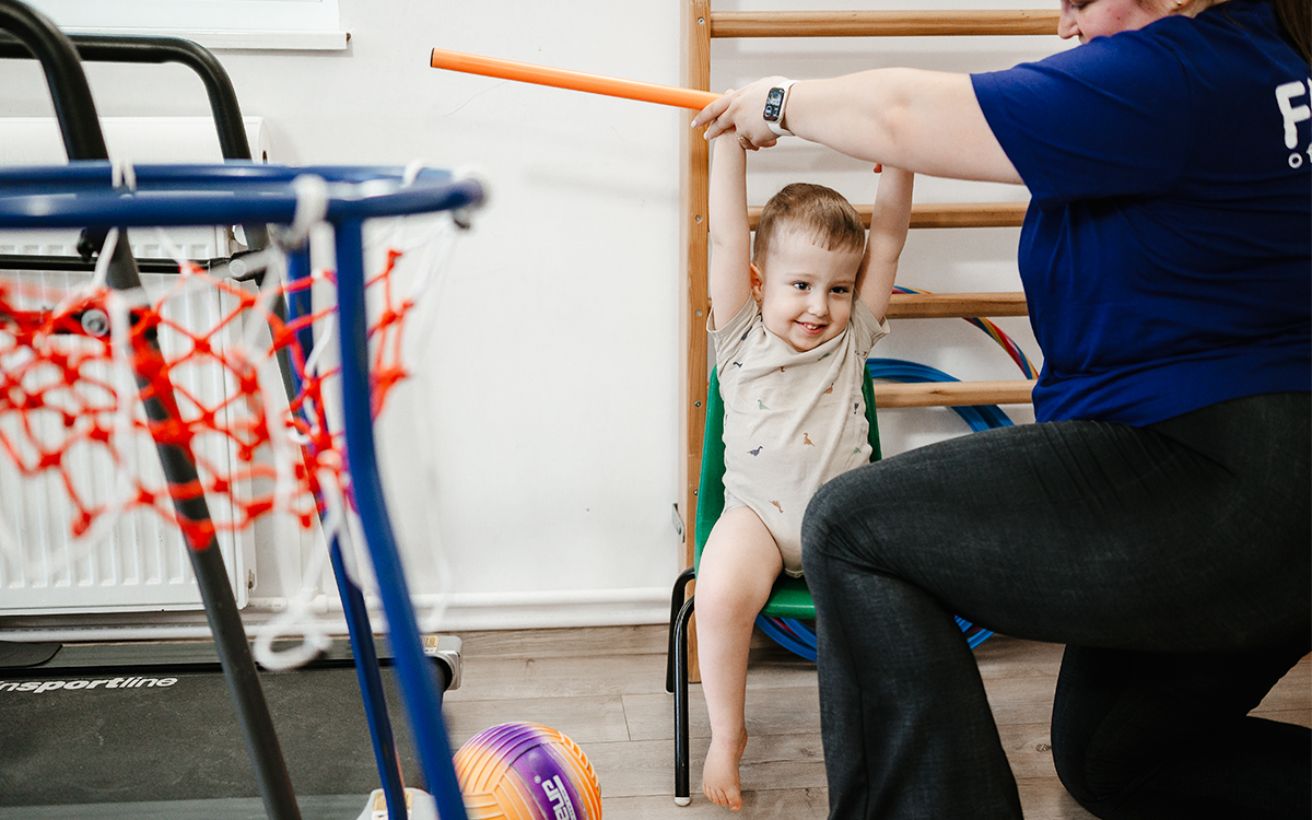 Photo of a young boy receiving physical therapy. He is reaching above his head for a pole the therapist is holding. He is smiling and wearing a beige babygrow