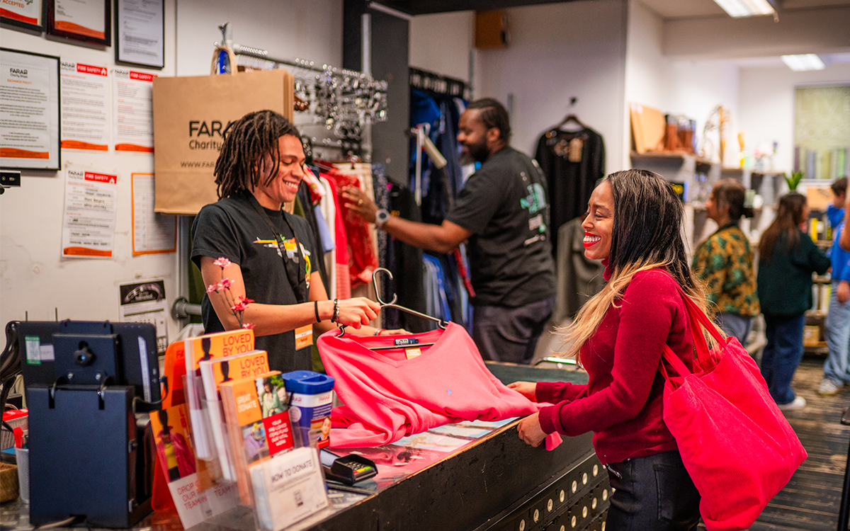 ChatGPT said: Customer smiling as a FARA shop volunteer serves her at the till.