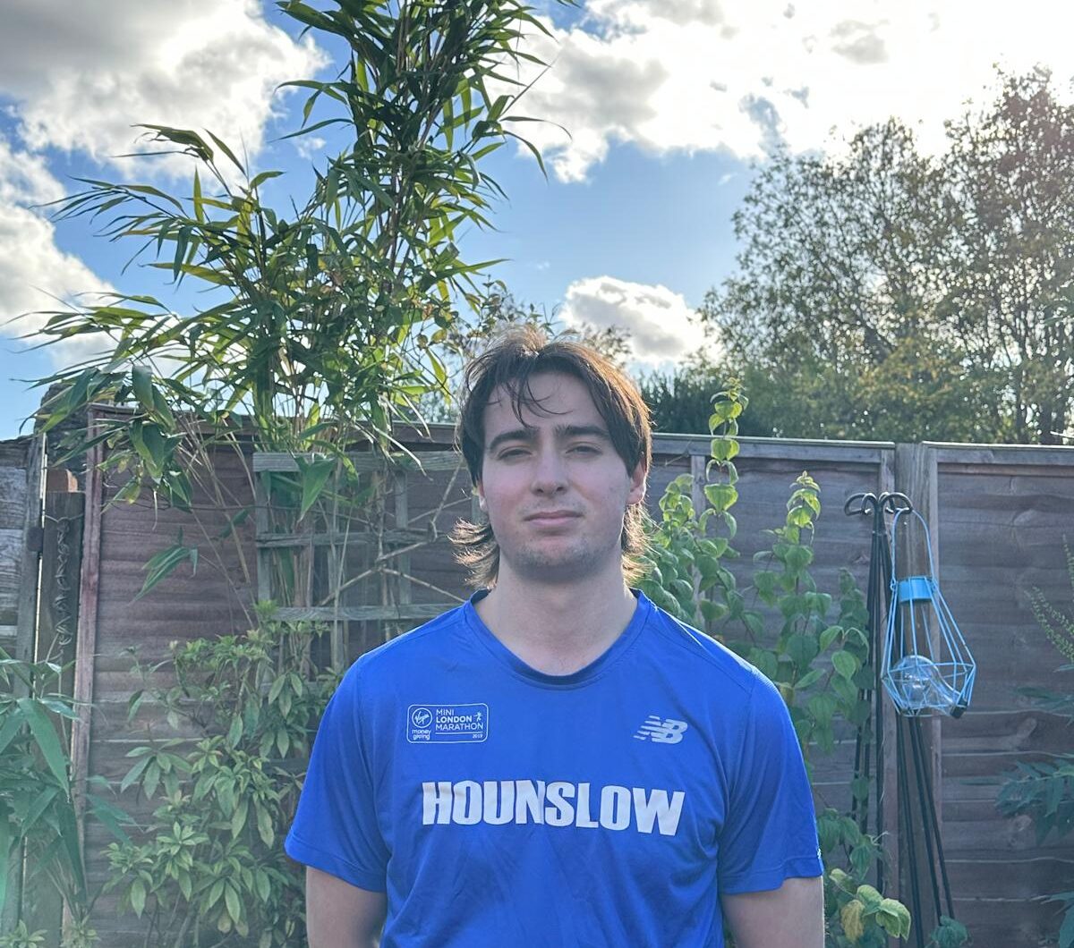 A photo showing a young man looking a the camera. He is in a garden and wearing a blue athletic top