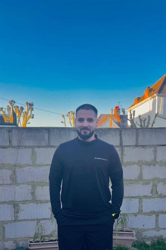 A photo of Florin-Maris standing in front of a breeze block wall with his hands in his pockets smiling directly at the camera. He has short brown hair and is wearing a navy jumper